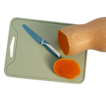 A light green rectangular cutting board with a handle cutout, shown on a white background with a butternut squash half, a carrot slice, and a blue-handled knife.