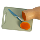 A light green rectangular cutting board with a handle cutout, shown on a white background with a butternut squash half, a carrot slice, and a blue-handled knife.