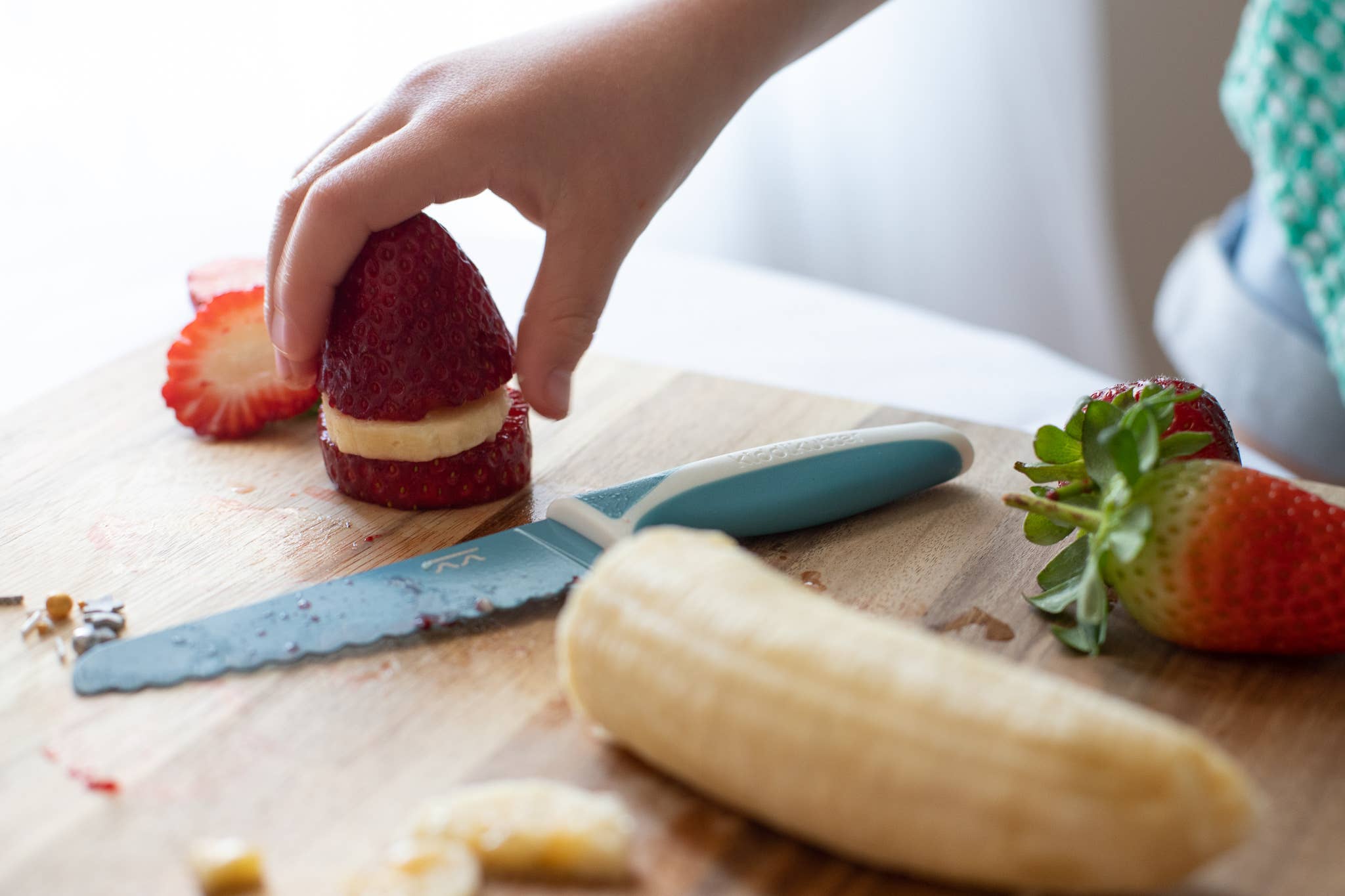 Person preparing a dessert with strawberries and banana on a wooden cutting board using KiddiKutter