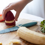 Person preparing a dessert with strawberries and banana on a wooden cutting board using KiddiKutter