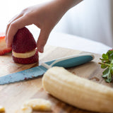 Person preparing a dessert with strawberries and banana on a wooden cutting board using KiddiKutter