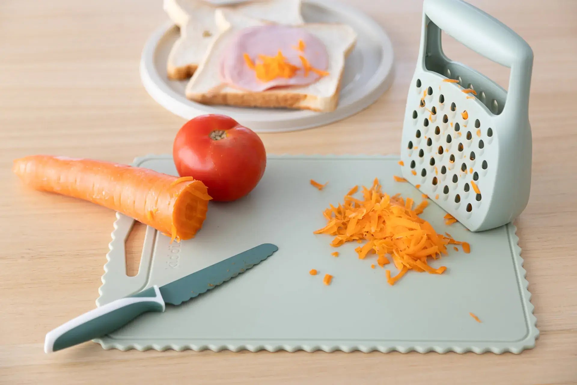 Chopping board with grated carrot, knife, tomato, and a plate of sandwich on a wooden surface.