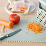Chopping board with grated carrot, knife, tomato, and a plate of sandwich on a wooden surface.