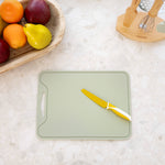 A sage green silicone cutting board with a handle cutout, shown on a light marble countertop with a yellow-handled knife resting on it. A wooden bowl of fruit and a wooden utensil holder are visible in the background.