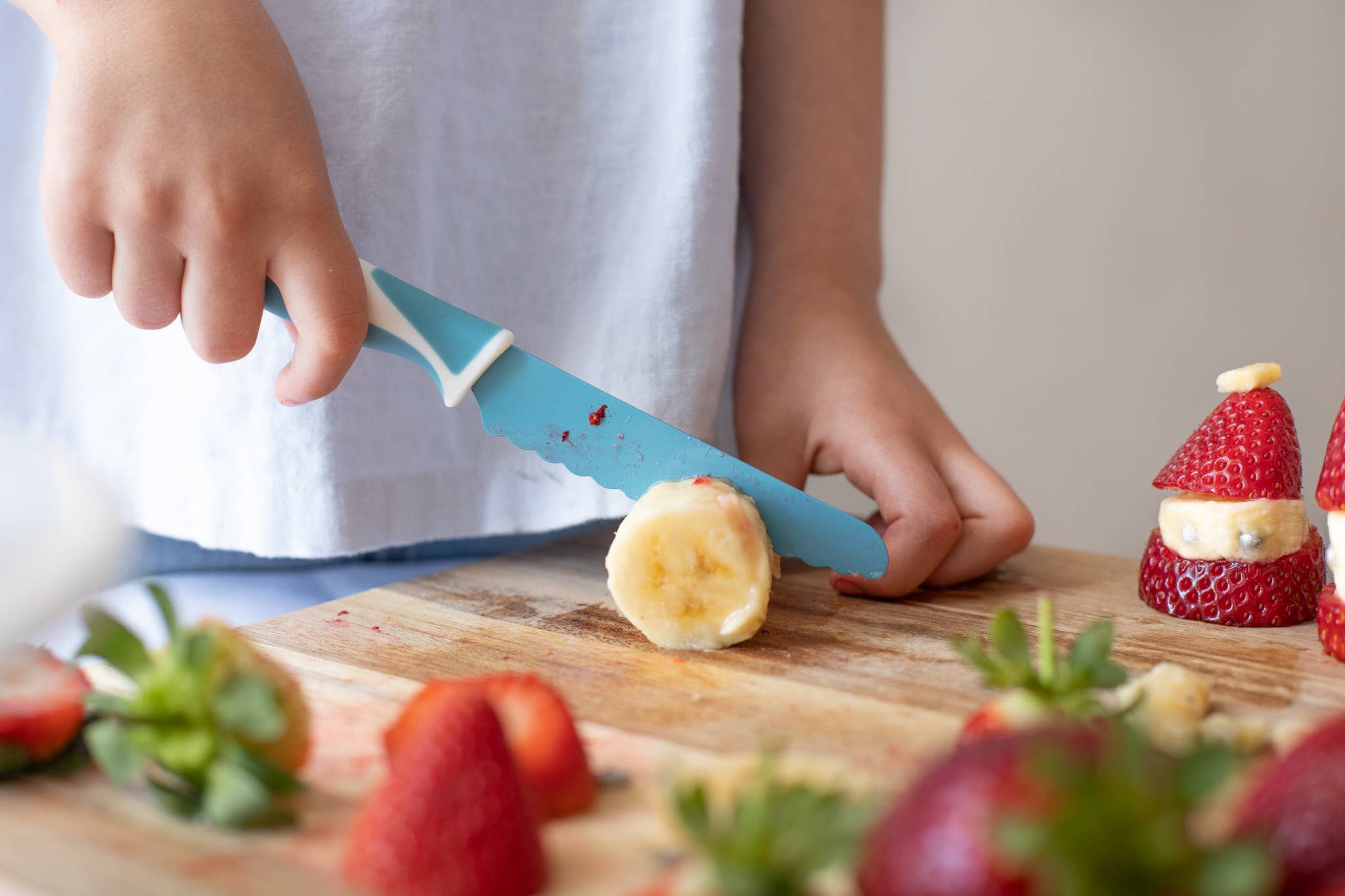 A person using a kiddikutter knife to cut a banana on a wooden cutting board with strawberries and a Santa Claus decoration in the background.