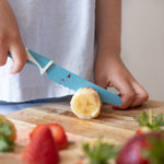 A person using a kiddikutter knife to cut a banana on a wooden cutting board with strawberries and a Santa Claus decoration in the background.
