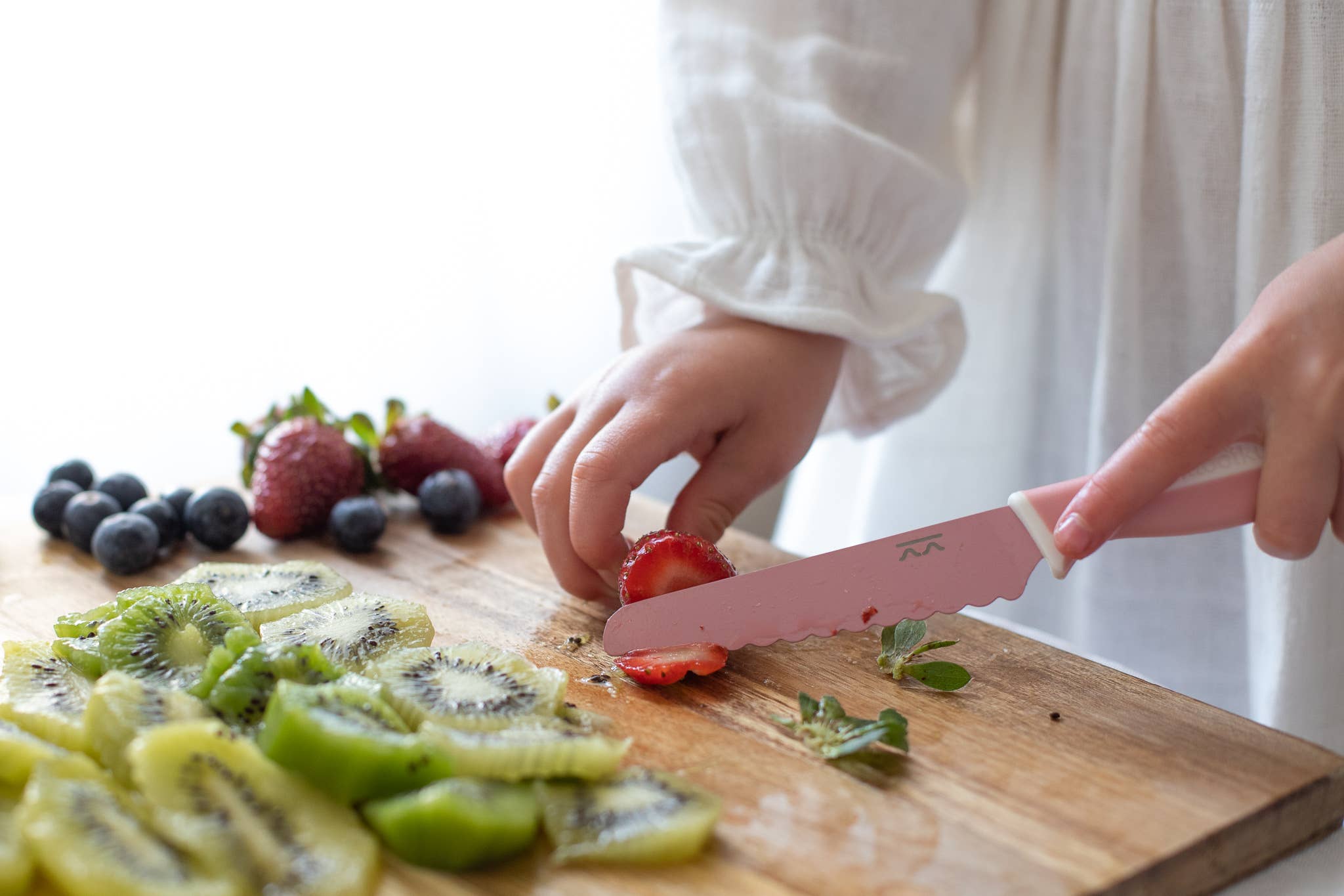 Person cutting strawberries with a pink knife on a wooden cutting board with kiwis and berries.