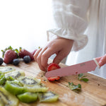 Person cutting strawberries with a pink knife on a wooden cutting board with kiwis and berries.