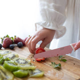 Person cutting strawberries with a pink knife on a wooden cutting board with kiwis and berries.
