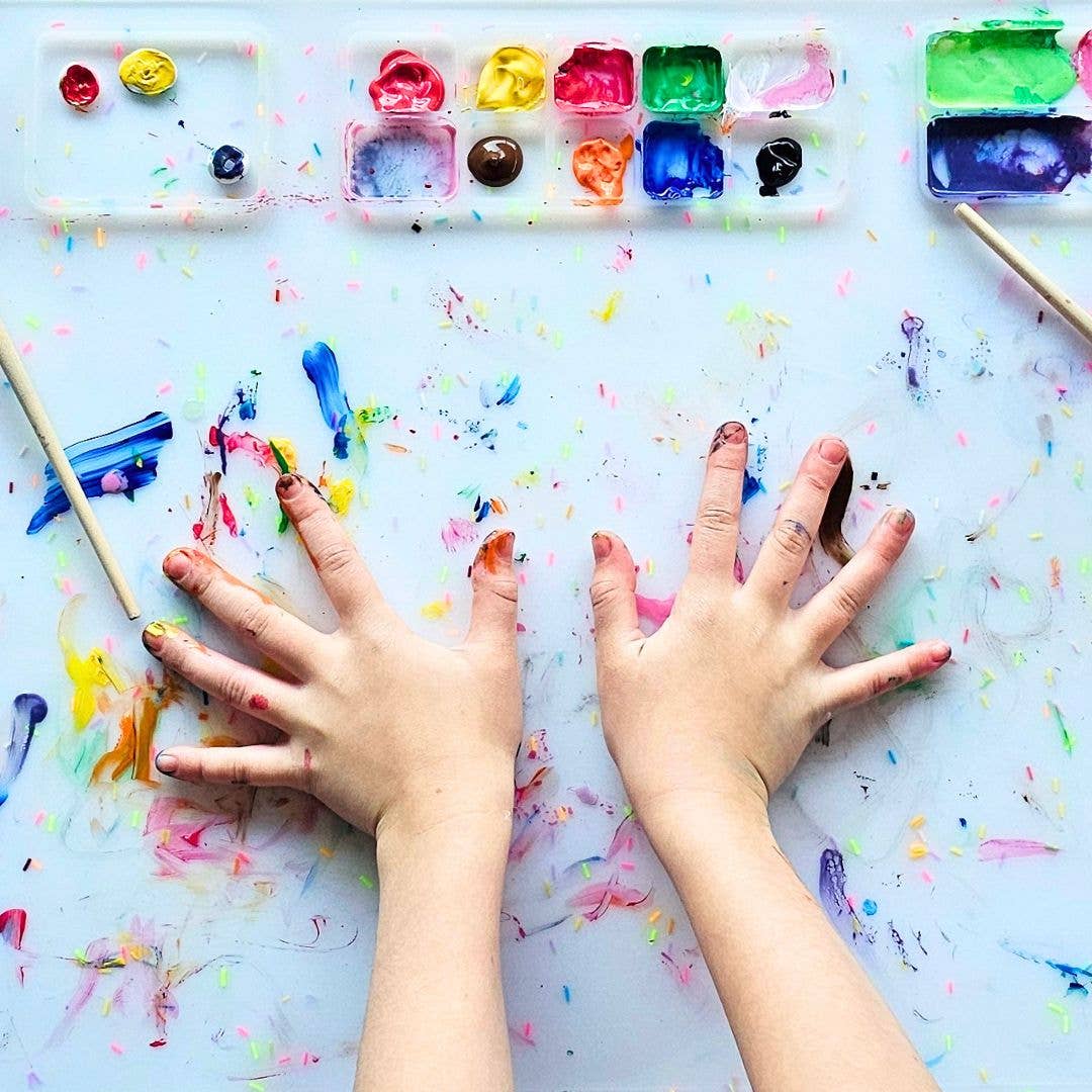 Two hands with paint splashes on a colorful painting surface with art supplies.