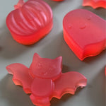 Red Halloween-themed jelly shaped like a bat, pumpkin, and ghost on a grey surface.