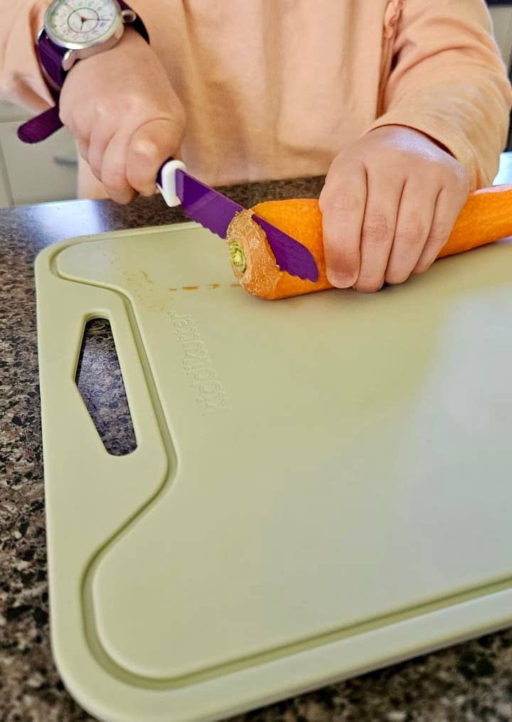 A person using a purple-handled vegetable peeler to remove the skin from an orange carrot on a light green cutting board, set on a dark granite countertop.