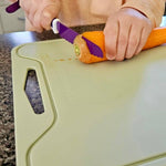 A person using a purple-handled vegetable peeler to remove the skin from an orange carrot on a light green cutting board, set on a dark granite countertop.