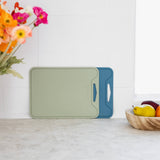Two rectangular cutting boards with integrated handles, one in sage green and one in blue, leaning against a white tiled kitchen wall on a light countertop. A vase of flowers and a bowl of fruit are partially visible in the scene.