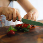 Child's hands using a kiddikutter knife to cut strawberries on a wooden cutting board.