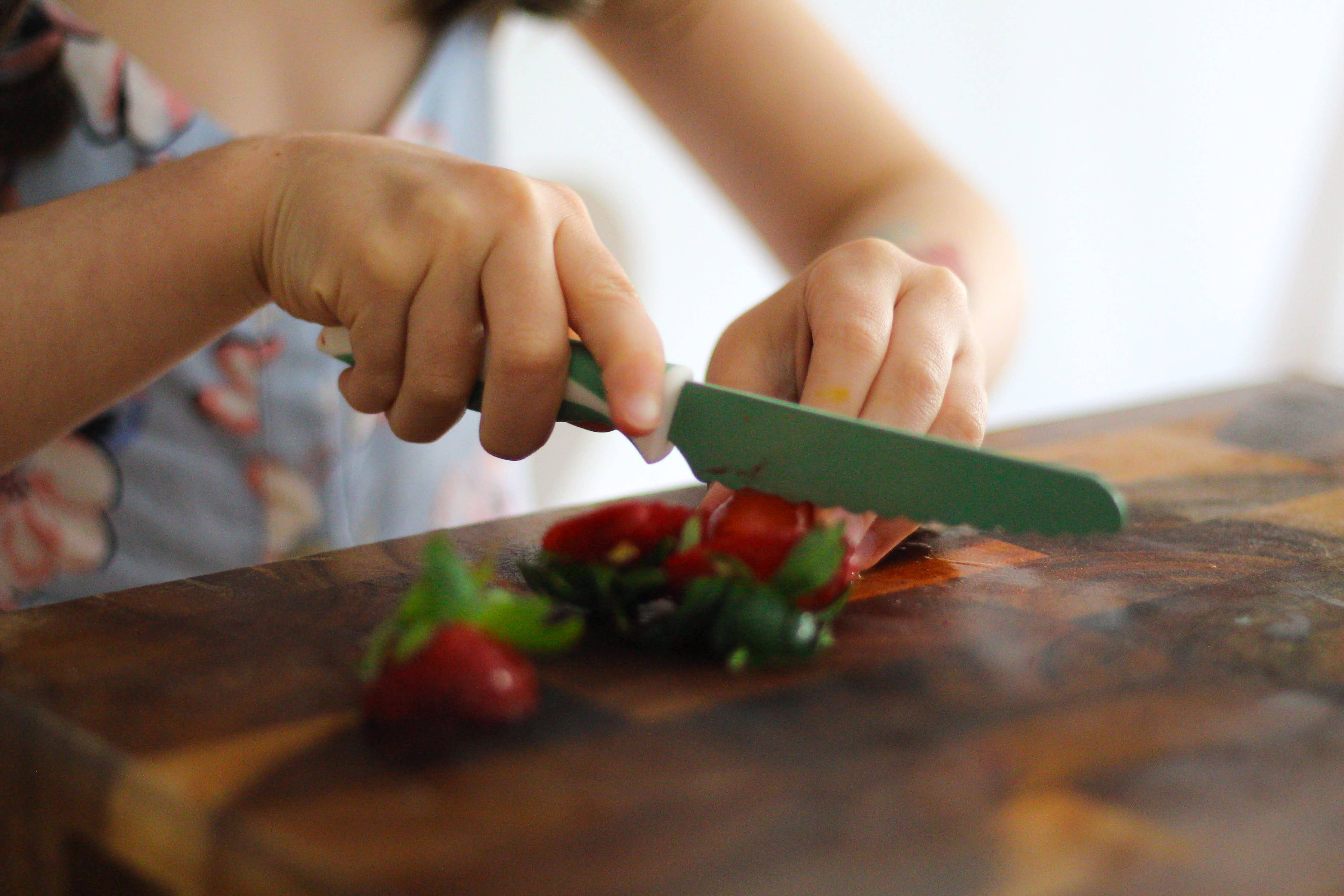 Child's hands using a kiddikutter knife to cut strawberries on a wooden cutting board.