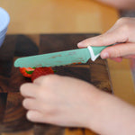 Person cutting a strawberry with a kiddikutter sea green knife on a wooden cutting board.