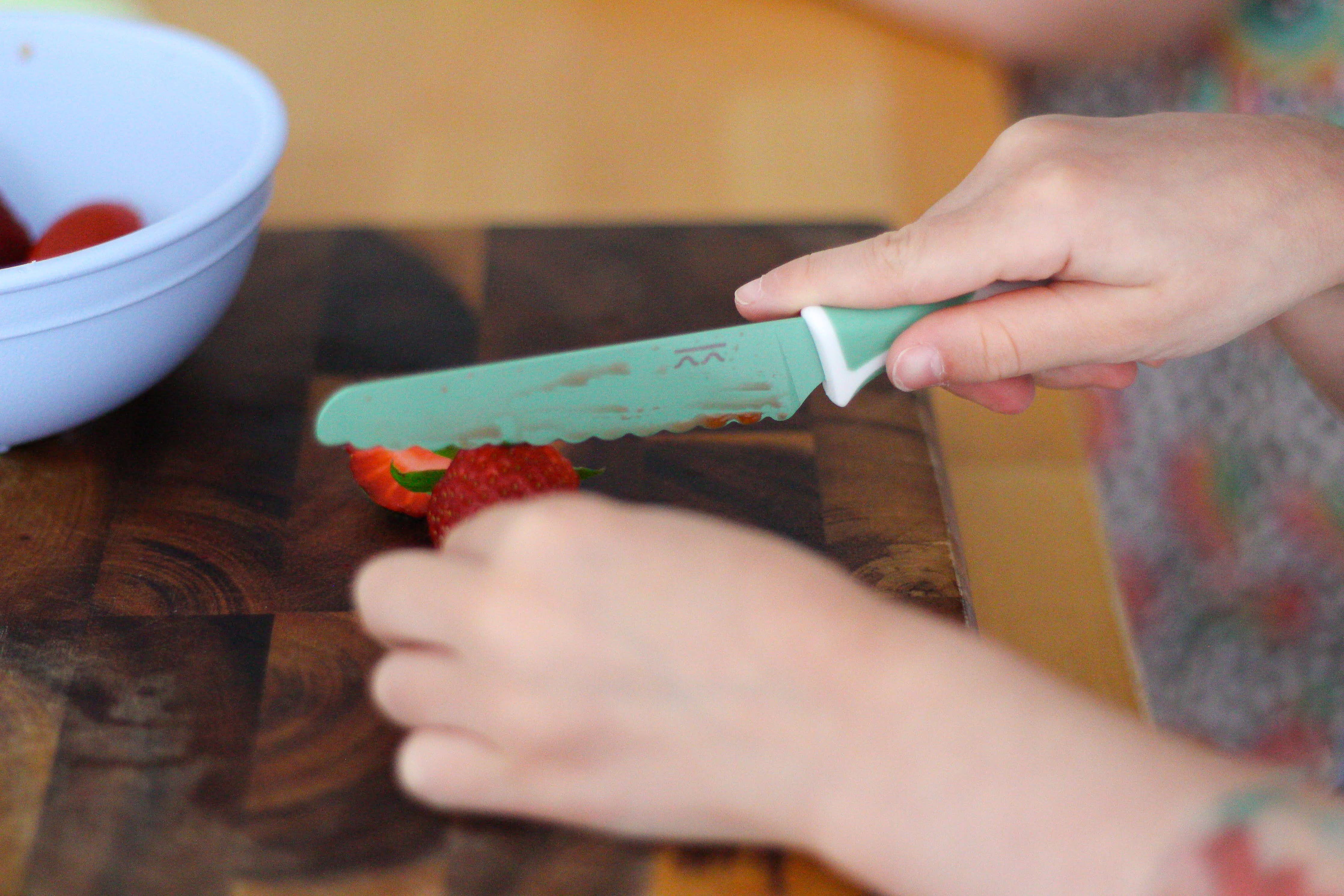 Person cutting a strawberry with a kiddikutter sea green knife on a wooden cutting board.